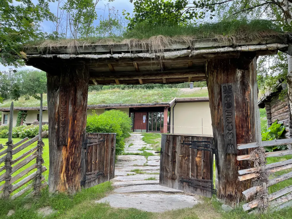 En åpen gammeldags port med tak, porten står på en brolagt sti som fører tl et avlangt grått hus i bakgrunnen