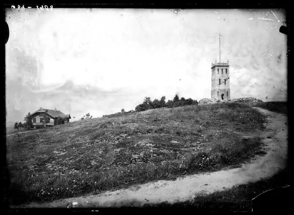 Sort-hvit-bilde som viser en gress-slette med en grusvei som går i en sving opp mot et tårn til høyre i bildet. Til venstre i bildet ser vi et hus i tre.