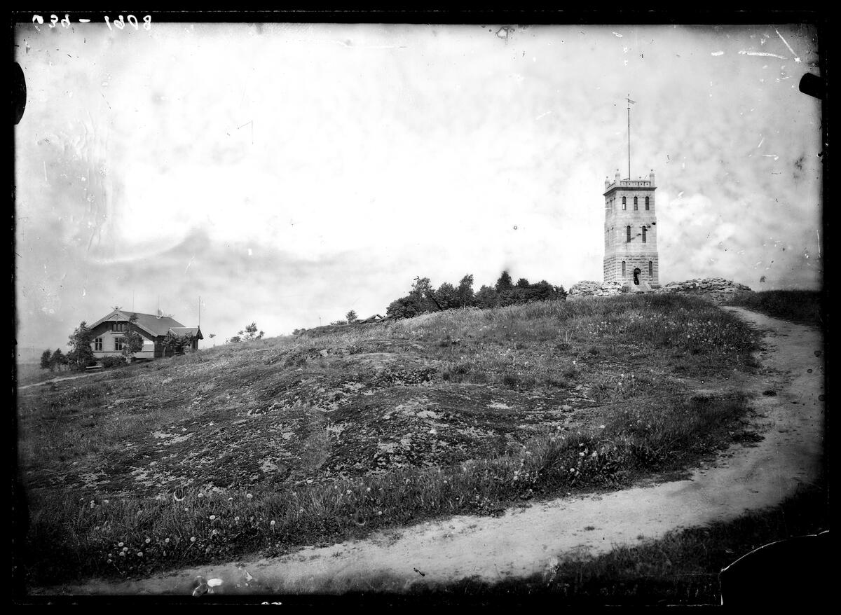 Sort-hvit-bilde som viser en gress-slette med en grusvei som går i en sving opp mot et tårn til høyre i bildet. Til venstre i bildet ser vi et hus i tre.