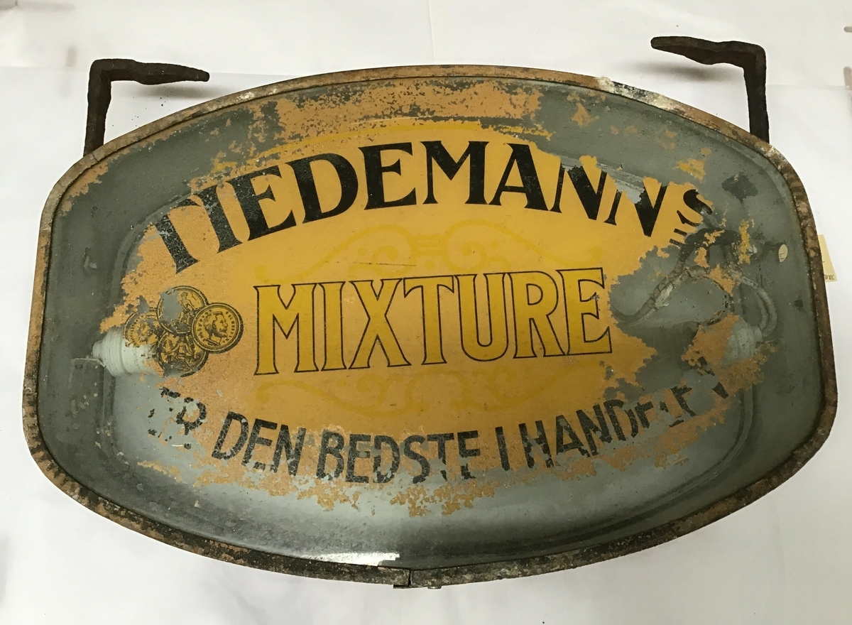 Reklameskilt for Tiedemanns tobakksvarer. Skiltet er avlang, ellipseforma men med skrå kortsider. Ulike motiv på sidene, det er opplegg for elektrisk lys med lyspærer  inne i skiltet.