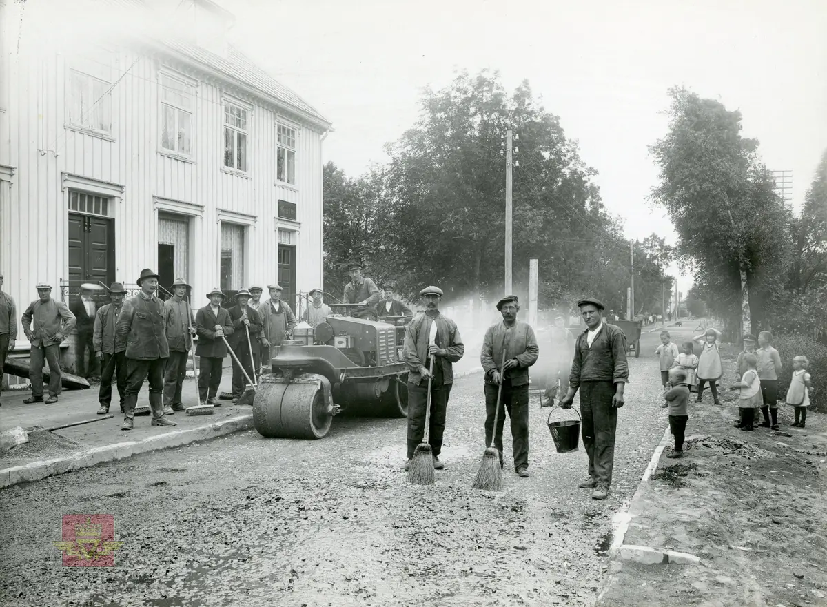 Prøveasfaltering i Kjøpmannsgata på Stjørdalshalsen 1923 - første asfaltering i dette fylke. To bilder.  Ene bildet har ramme  med håndskrevet tekst. 
Mannen med hatt og leggings er oppsynsmann Aasen. Barn står og ser på. 
Det var mange mann i arbeid for å få asfalten utover gata. Ingen tvil om hvem som er sjefen her.