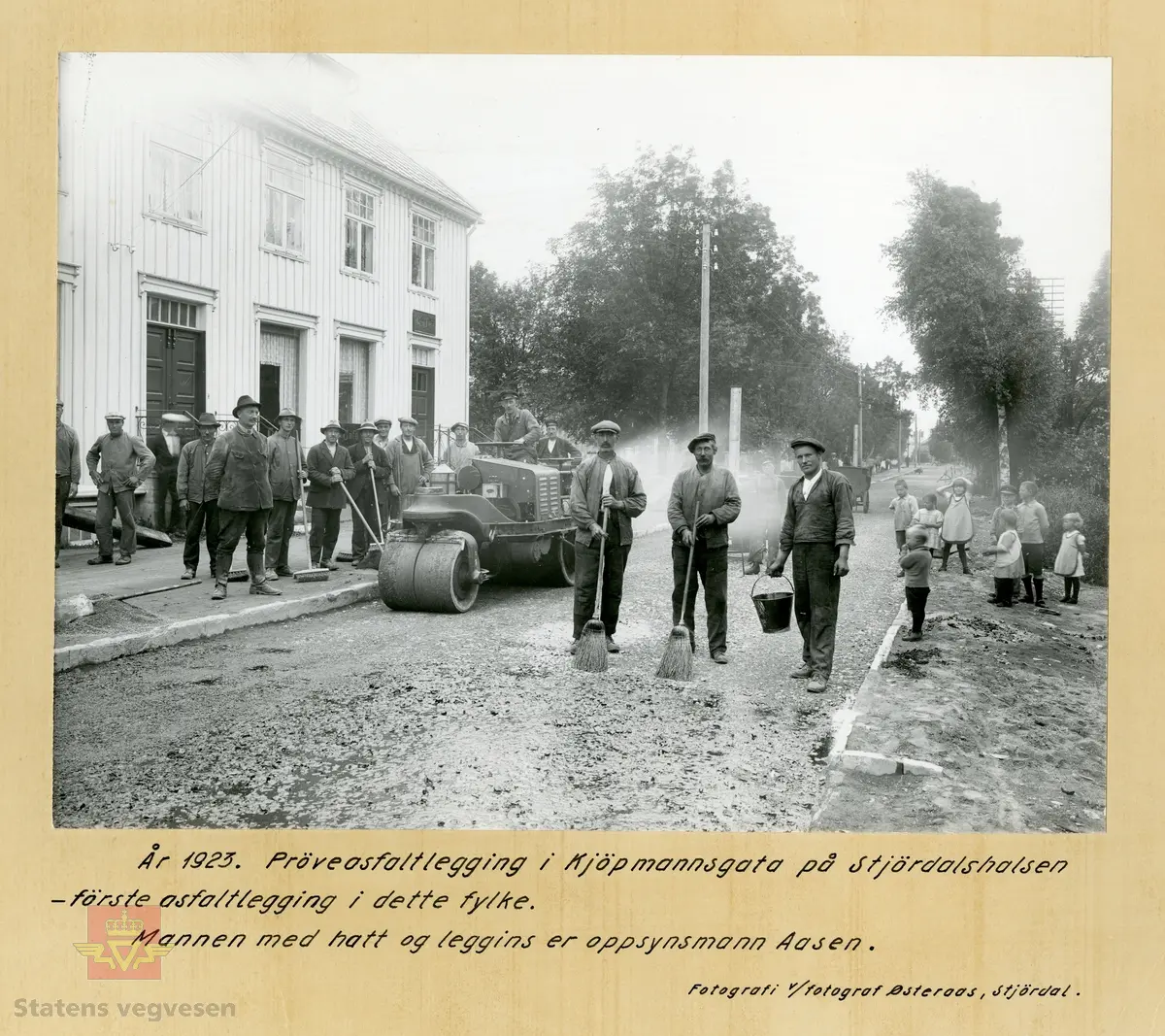 Prøveasfaltering i Kjøpmannsgata på Stjørdalshalsen 1923 - første asfaltering i dette fylke. To bilder.  Ene bildet har ramme  med håndskrevet tekst. 
Mannen med hatt og leggings er oppsynsmann Aasen. Barn står og ser på. 
Det var mange mann i arbeid for å få asfalten utover gata. Ingen tvil om hvem som er sjefen her.