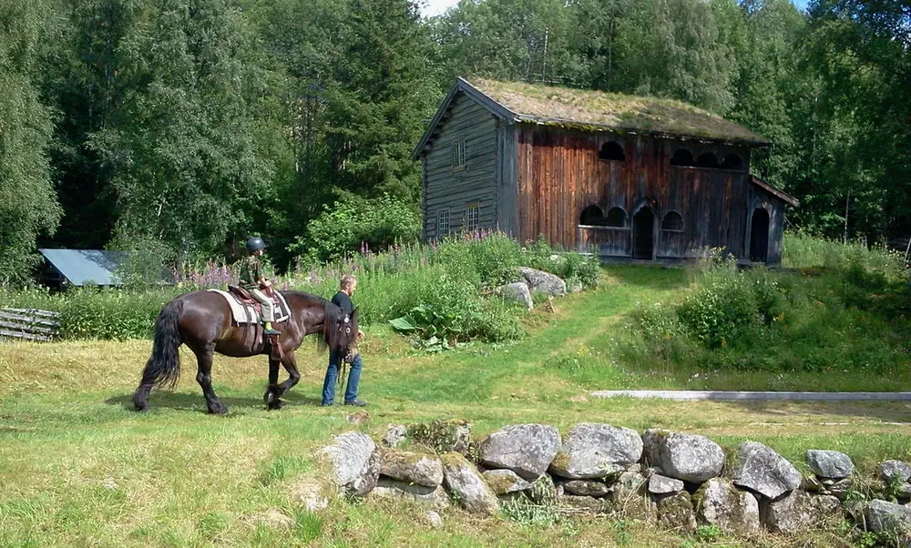 En mann som leier en hest med et barn på ryggen ned en sti, på en bakke bakenfor menneskene står et mellomstort brunt hus med gress på taket, bakgrunnen er fylt med trær