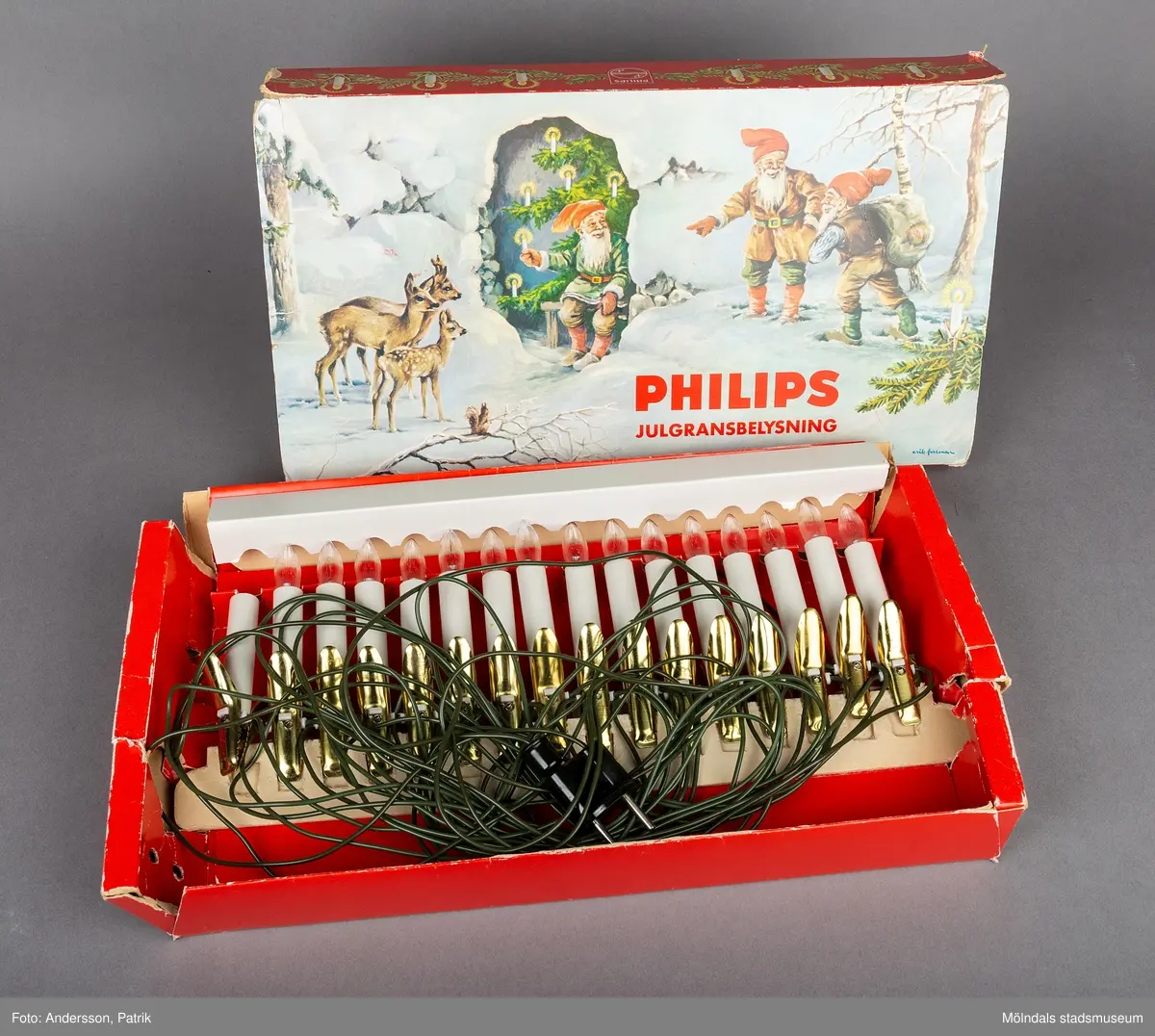 En papplåda som innehåller en elektrisk julgransbelysning av märket Philips. Lådans utsida har en målad vinterbild som föreställer ett vinterlandskap med tre tomtar ute i snön. I mitten på bilden finns en julgran med belysning. Till vänster på bilden, står två rådjur och tittar på tomtarna. 
På lådans baksida finns en monteringsanvisning. Nedtill finns en signatur som anger att det Erik Forsman som har målat bilden.
I lådan finns en elektrisk ljusslinga med 16 ljus. Det finns även en tiilhörande plastpåse med 15 glödlampor.
Julgransbelysningen är inköpt 1965 av Gerd och Jan Kilnäs som bodde i Kallebäck. Belysningen är inköpt på Kallebäcks Livs den 23 december 1965. Gerd och Jan Kilnäs flyttade till Mölnlycke år 1968 och har använt ljusslingan varje år fram till 2014, då togs den över av Maria Kilnäs.
Elektriska julgransbelysningar började tillverkas i slutet på 1800-talet. Det var först under mellankrigstiden som den började användas i större utsträckning.
Innan det använde man stearinljus.
