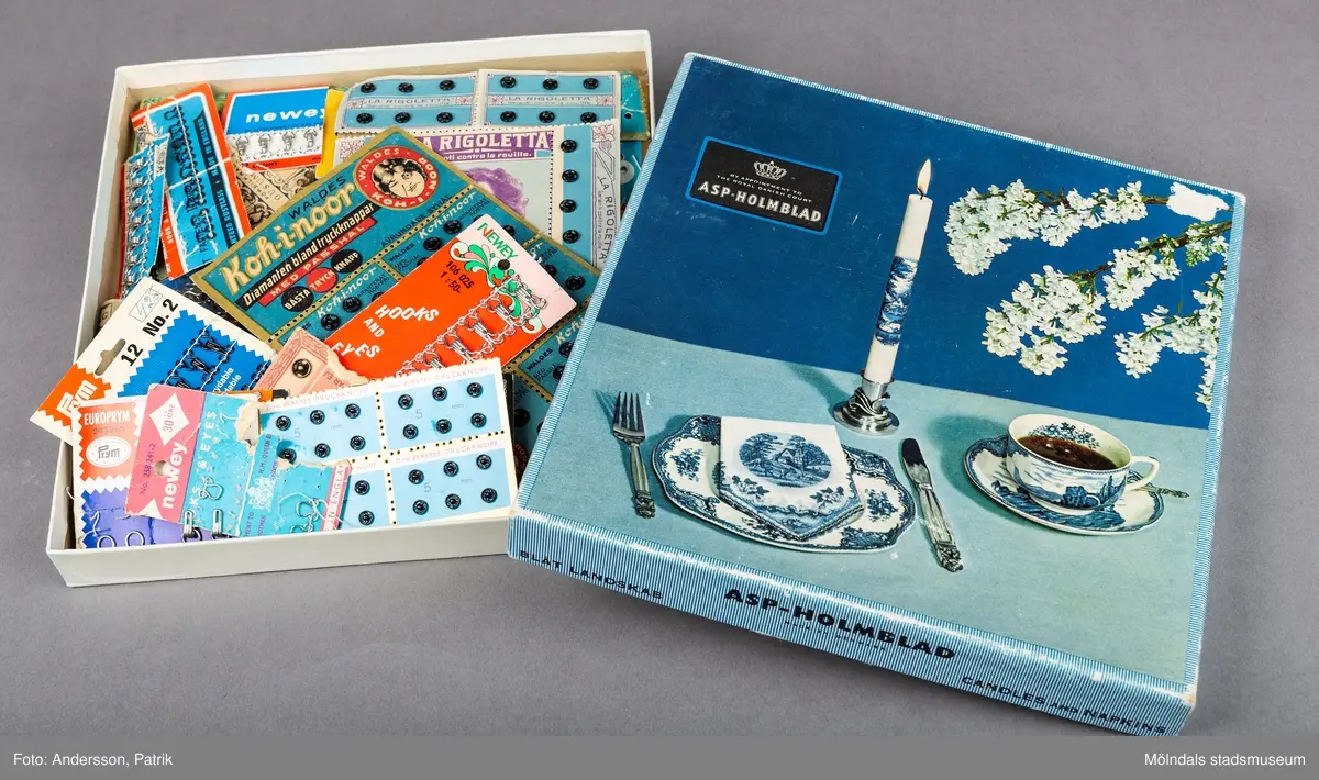 En blå kvatratisk pappask som ursprungligen innehållit ljus och servetter. Asken ser ut att vara en presentask. På framsidan finns en bild som föreställer ett dukat bord. På bilden finns ett kaffekuvert bestående av en kopp, en tallrik med bestick och ett ljus i en silverljusstake. På höger sida finns en vit syrenkvist.
På askens sida står det Blåt Landskap.
Asken innehåller en stor mängd sytillbehör i form av små knappar och hyskor i metall.