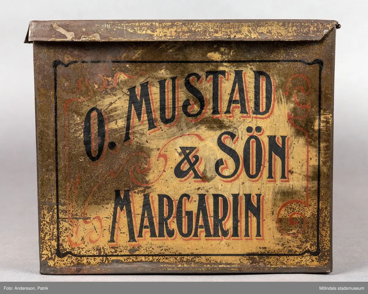 En plåtlåda av större format som innehållit Margarin. Den rektangulära lådan är gjord av plåt. Lådan är brungrå till färgen och är repad och skavd, men texten är välbevarad. På locket finns texten O.MUSTAD&SÖN MARGARIN skriven med svart text.
I mitten på locket finns en utsmyckning i form av en blomma och en girlang i orange färg. Bilden och texten är inramad av en snirklig ram i svart färg. 
På lådans långsidor står det även Mölndal med stora bokstäver.
Mustads margarinfabrik var verksam i Mölndal under åren 1897-1960.
Fabriken var belägen vid Stensjön.