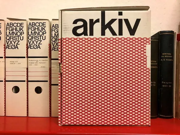 Bildet viser en rød hylle der vi ser ryggene til tre bokser med alfabetet, noen linjer og et hull på baksiden. En av boksene er oppstilt slik at vi ser siden av boksen. Øverst står det skrevet arkiv. Nedenfor er det rødt rutemønster med hvit bakgrunn.