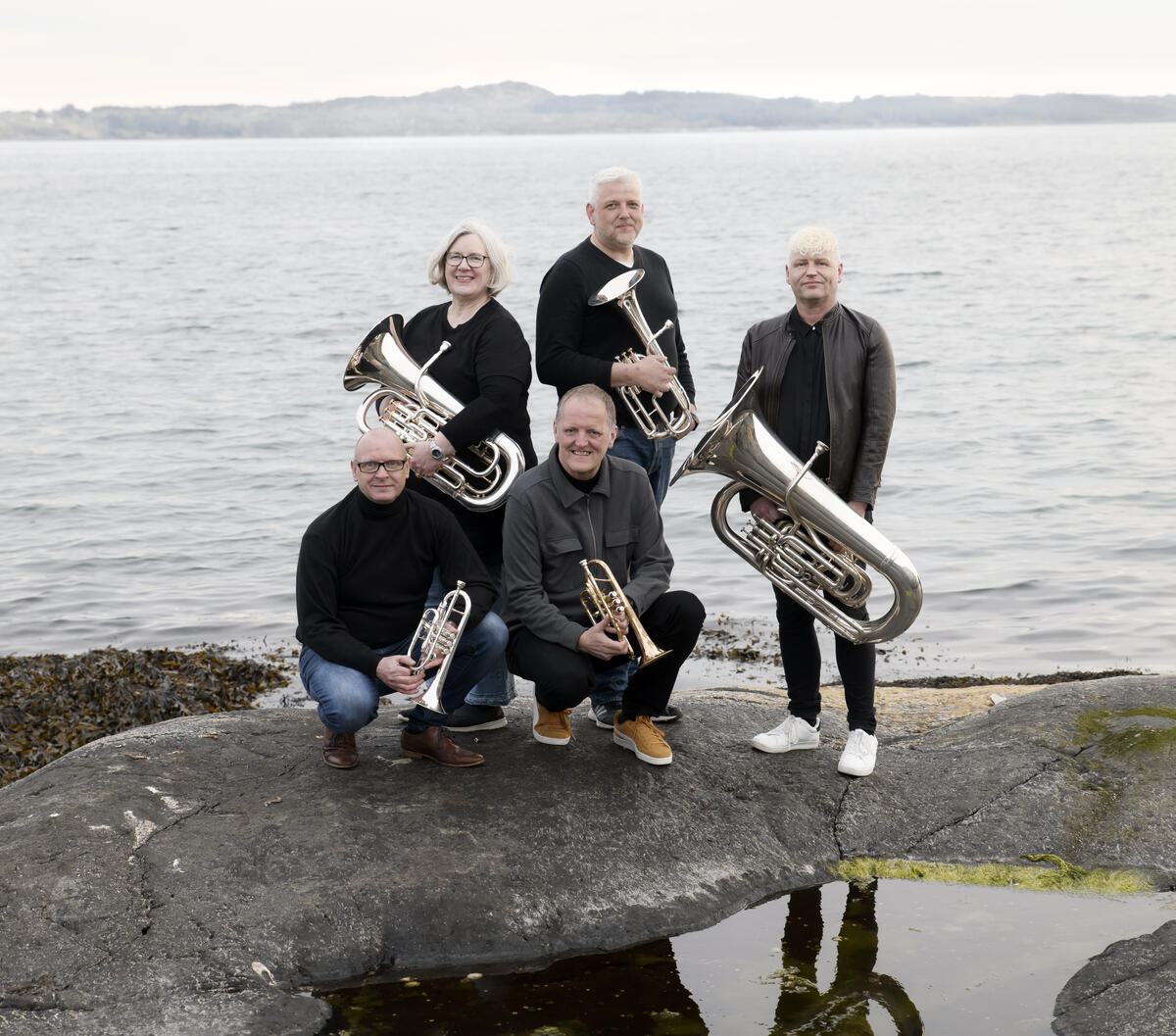 Fem brassmusikanter med instrumenter på svaberg i sjøkanten
