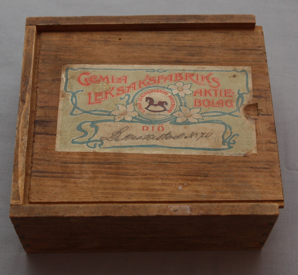 Bygglåda, rektangulär låda av furu med inskjutbart lock från Gemla Leksaksfabriks Aktiebolag (1866-1884). Utan leksaker innuti. Etikett på locket som föreställer en logga med en gunghäst omgiven av tre vita blommor. Etikettens yttre kanter har ett dekorelement föreställande ett slingrande band.