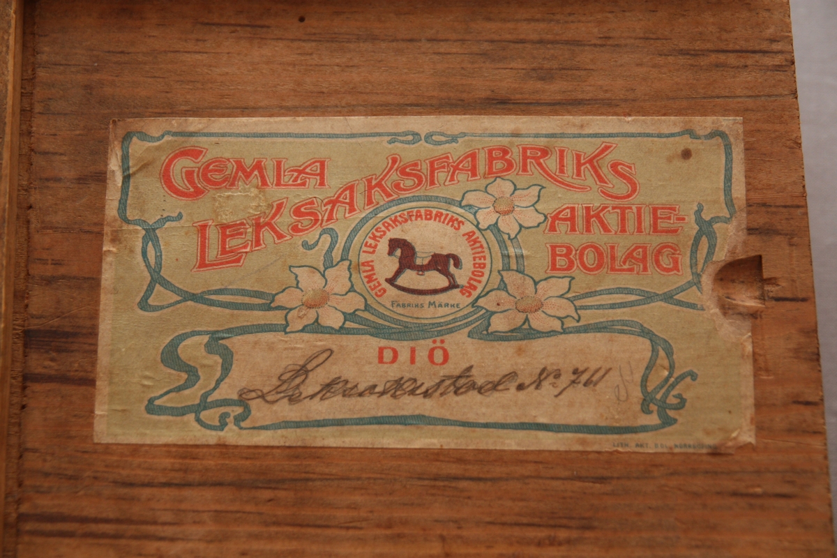 Bygglåda, rektangulär låda av furu med inskjutbart lock från Gemla Leksaksfabriks Aktiebolag (1866-1884). Utan leksaker innuti. Etikett på locket som föreställer en logga med en gunghäst omgiven av tre vita blommor. Etikettens yttre kanter har ett dekorelement föreställande ett slingrande band.