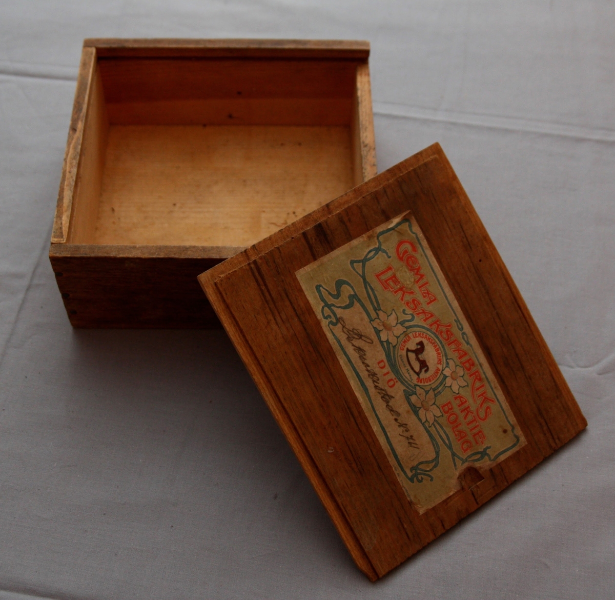 Bygglåda, rektangulär låda av furu med inskjutbart lock från Gemla Leksaksfabriks Aktiebolag (1866-1884). Utan leksaker innuti. Etikett på locket som föreställer en logga med en gunghäst omgiven av tre vita blommor. Etikettens yttre kanter har ett dekorelement föreställande ett slingrande band.