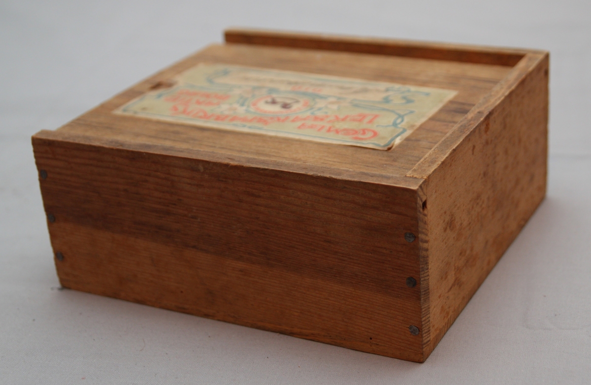 Bygglåda, rektangulär låda av furu med inskjutbart lock från Gemla Leksaksfabriks Aktiebolag (1866-1884). Utan leksaker innuti. Etikett på locket som föreställer en logga med en gunghäst omgiven av tre vita blommor. Etikettens yttre kanter har ett dekorelement föreställande ett slingrande band.