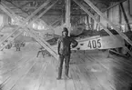 En militär flygare står i F-hangaren på Flygkompaniets verkstäder på Malmen, omkring 1920. I bakgrunden står flygplan Albatros och Morane-Saulnier MS 3L.