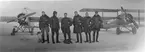 Grupporträtt. Sex stycken militära flygare och två hundar framför flygplan Thulin K och Triplanet på flygfältet på Flygkompaniet på Malmen, vintertid, 1918. Från vänster: Gösta D:son Carlson, Gustaf von Segebaden, Nils Rodéhn, Paul af Uhr, flygspanare B. T. Kjelldahl, Fredrik Adilz.