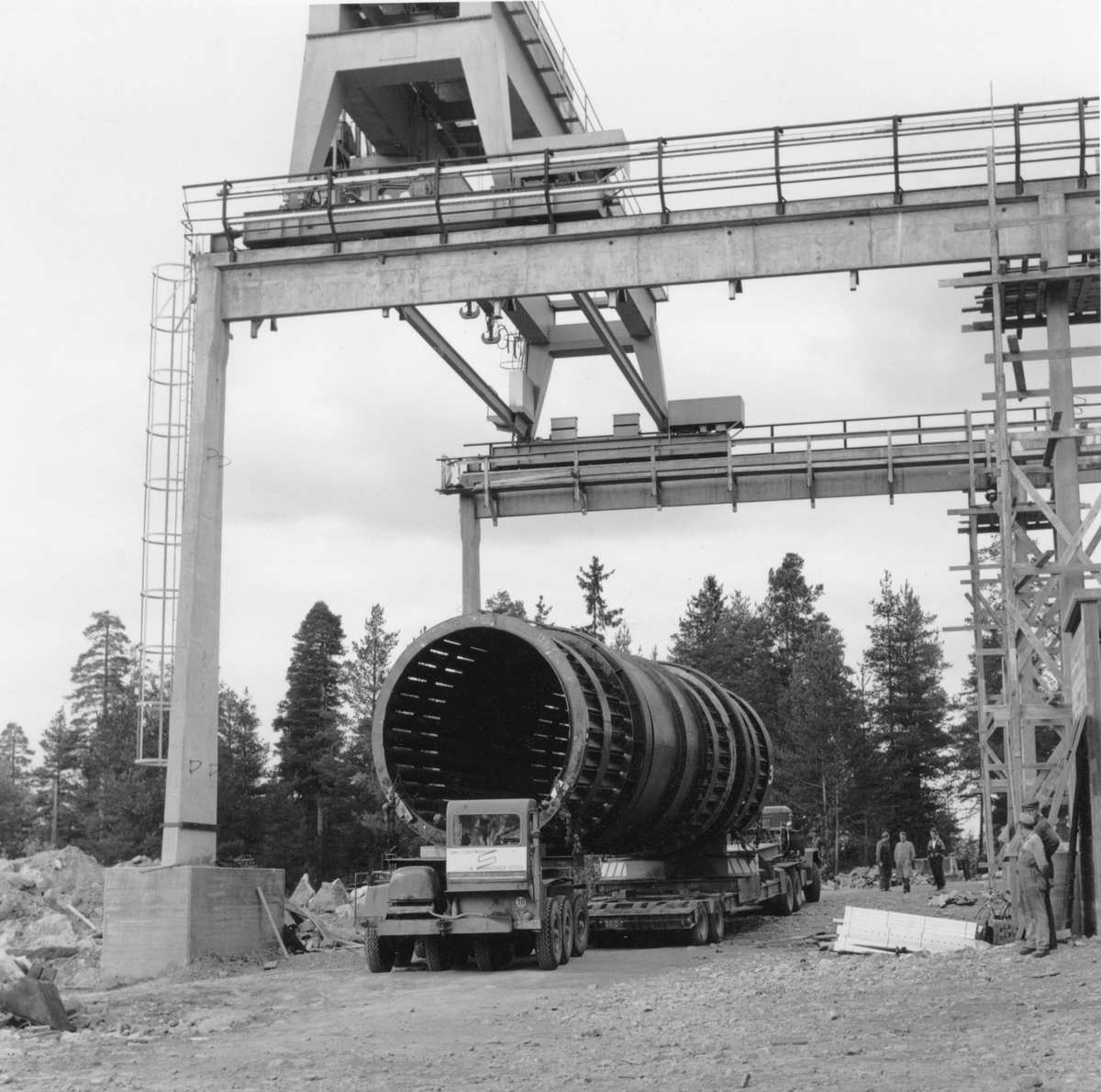 Transport av barketrommel til Geithus Bruk på Modum i Buskerud i 1966. Trommelen ble produsert ved A/S Myrens Verksted i Oslo.  På grunn av jernbaneunderganger og akseltrykkrestriksjoner kunne ikke trommelen transporteres hele vegen med lastebil.  Den ble derfor delt i to deler, hver med cirka 11 meters lengde, som ble transportert til Drammen med lastebåt.  Derfra ble det brukt lastebil, delvis om natta for å unngå å skape hindringer for annen trafikk.  Dette fotografiet er tatt i dagslys på et tidspunkt da to lastebiler som bar hver ende av en halv barketrommel sto parkert på det nye industriområdet, under den nybygde kranbanen, som antakelig skulle løfte trommelen videre moot monteringsstedet. En del karer hadde samlet seg for å se den sjeldent store og tunge transporten.