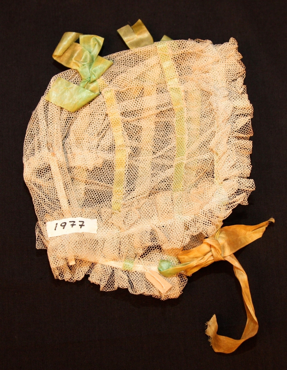 Dopmössa i tyll med rosetter av sidenband. Tyllen sydd i volanger kring kanterna.