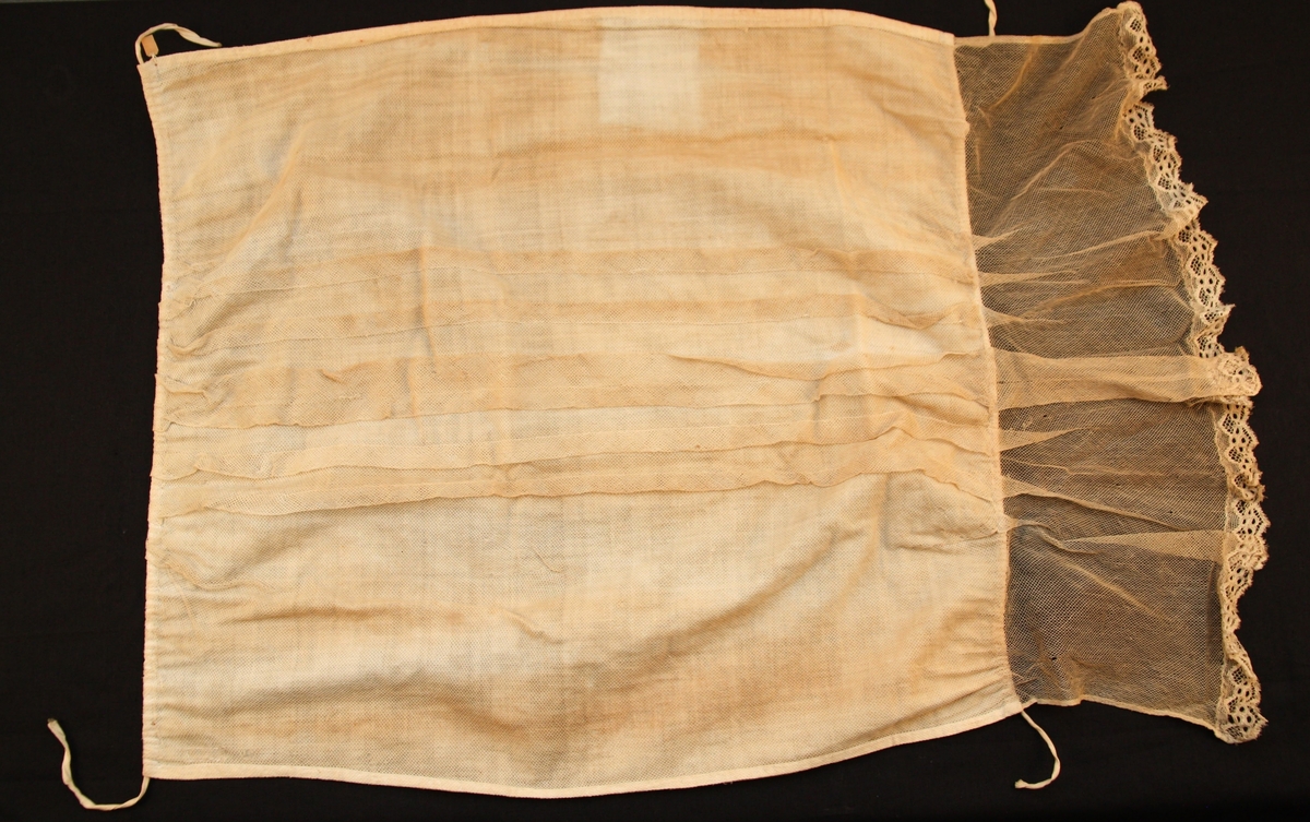 Ett fyrsidigt vitt tygstycke vars främre sida är överdraget med vit tyll. I tygets nedre kant finns en spetsbård. Längs mitten har tyllen sytts i dekorativa veck.