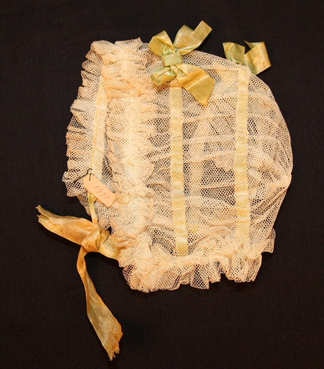 Dopmössa i tyll med rosetter av sidenband. Tyllen sydd i volanger kring kanterna.