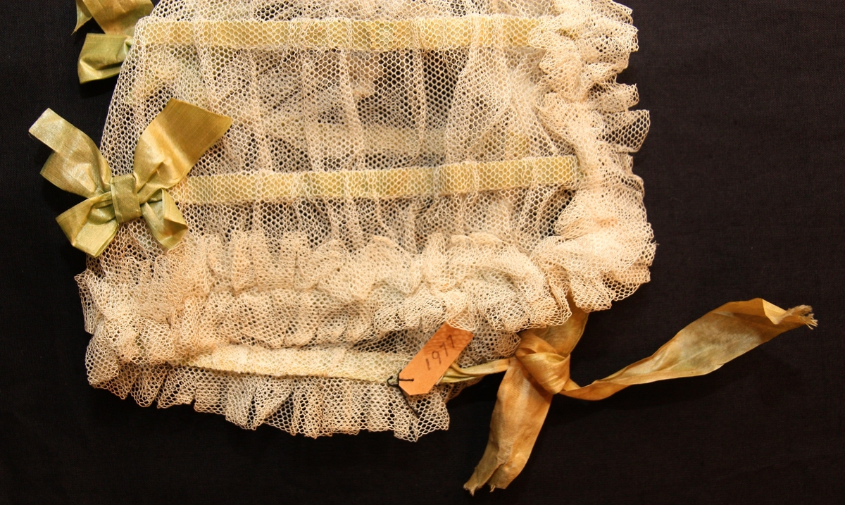 Dopmössa i tyll med rosetter av sidenband. Tyllen sydd i volanger kring kanterna.