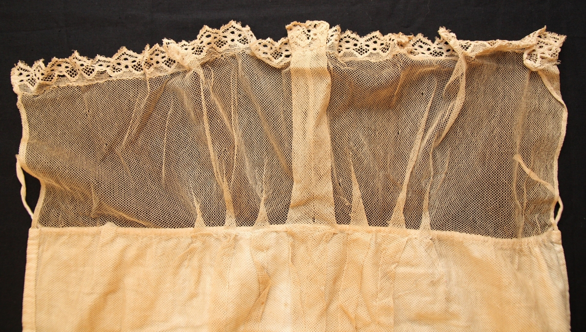 Ett fyrsidigt vitt tygstycke vars främre sida är överdraget med vit tyll. I tygets nedre kant finns en spetsbård. Längs mitten har tyllen sytts i dekorativa veck.