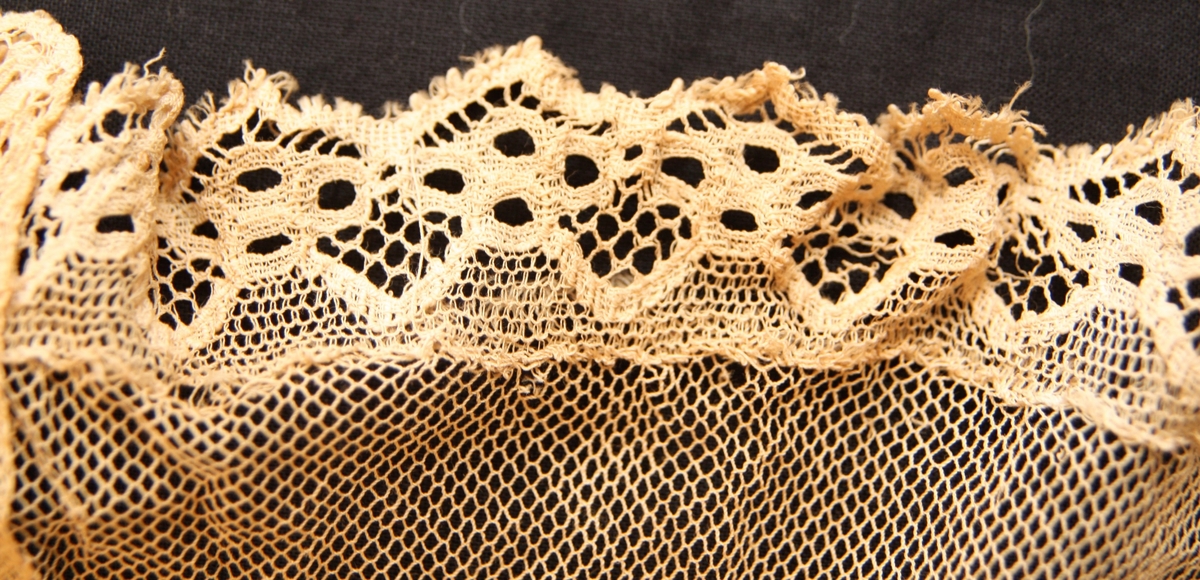 Ett fyrsidigt vitt tygstycke vars främre sida är överdraget med vit tyll. I tygets nedre kant finns en spetsbård. Längs mitten har tyllen sytts i dekorativa veck.