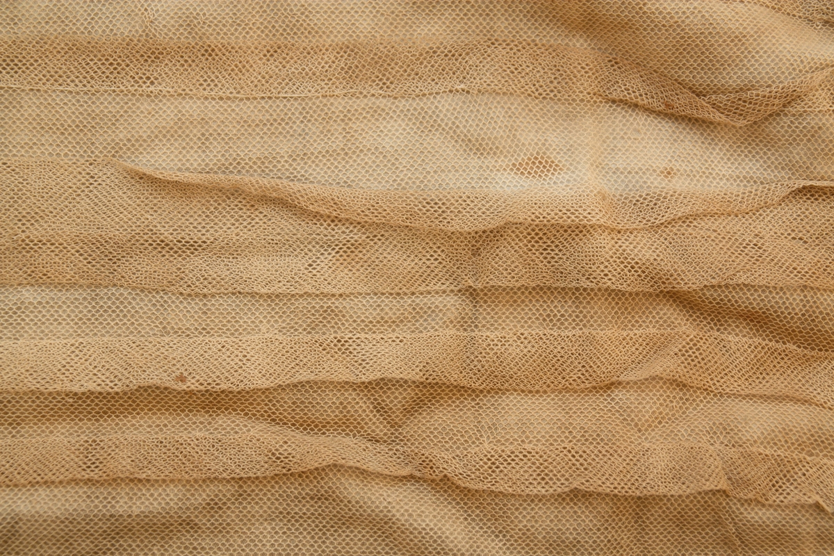 Ett fyrsidigt vitt tygstycke vars främre sida är överdraget med vit tyll. I tygets nedre kant finns en spetsbård. Längs mitten har tyllen sytts i dekorativa veck.