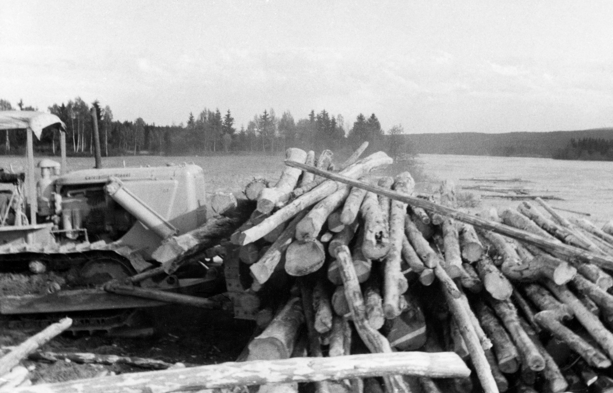 Utislag av fløtingstømmer i Glomma ved hjelp av bulldoser.  Prosessen er fotografert fra sida.  Vi ser fronten på bulldoseren, som skyver en haug med handbarket tømmer utfor elvebakken ved hjelp av skjæret.  I bakgrunnen til høyre ses elva, der det flyter tømmer langs det landet der utislaget foregår.  Bakgrunnen i høyre del av bildet består av ei flat, vegetasjonsløs slette (muligens en uspiret åker) med en bakenforliggende skog. Utislag med maskinelle hjelpemidler var noe nytt i 1950-åra.