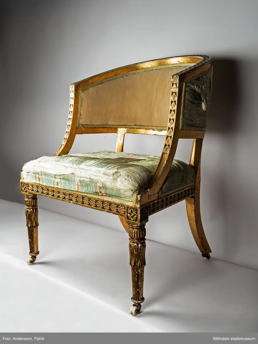 Fåtölj, s.k. "baljfåtölj", sengustaviansk, tillverkad omkring år 1800. 

Kraftigt svängd rygg, raka sarger med skuren dekor, svarvade, bladhölstrade och kannelerade framben, sabelformade bakben. 
Fast stoppad sits och rygg, med sliten, senare klädsel i grönrandigt siden. Bevarad originalstoppning.

Senare bemålning i guld. Äldre rödbrun färg kan anas under förgyllningen.
Benen har kortats av och kompletterats med trissor (hjul) i mässing under 1800-talets andra hälft.

Sitthöjd 440 mm, Bredd 610 mm.
Material: Björk, Tageltyg, Sadelgjord.

Fåtöljen är en av ett par. Båda är signerade med brännstämpel AIS på den bakre sargen. Det finns fyra kända snickare i Lindome som använde signaturen AIS – Anders Johansson i Hassungared (1780-1847), Anders Jonsson i Ingemantorp (1770-1832), Anders Jönsson i Gastorp (1778-1858) och Andreas Johansson i Skår (1793-1871).

Inköpt på Kungsbacka Auktionskammare.
Samhör med MM05442