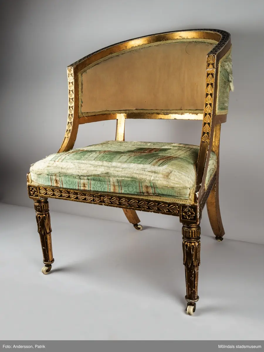 Fåtölj, s.k. "baljfåtölj", sengustaviansk, tillverkad omkring år 1800. 

Kraftigt svängd rygg, raka sarger med skuren dekor, svarvade, bladhölstrade och kannelerade framben, sabelformade bakben. 
Fast stoppad sits och rygg, med sliten, senare klädsel i grönrandigt siden. Bevarad originalstoppning.

Senare bemålning i guld. Äldre rödbrun färg kan anas under förgyllningen.
Benen har kortats av och kompletterats med trissor (hjul) i mässing under 1800-talets andra hälft.

Sitthöjd 440 mm, Bredd 610 mm.
Material: Björk, Tageltyg, Sadelgjord.

Fåtöljen är en av ett par. Båda är signerade med brännstämpel AIS på den bakre sargen. Det finns fyra kända snickare i Lindome som använde signaturen AIS – Anders Johansson i Hassungared (1780-1847), Anders Jonsson i Ingemantorp (1770-1832), Anders Jönsson i Gastorp (1778-1858) och Andreas Johansson i Skår (1793-1871).

Inköpt på Kungsbacka Auktionskammare.
Samhör med MM05442