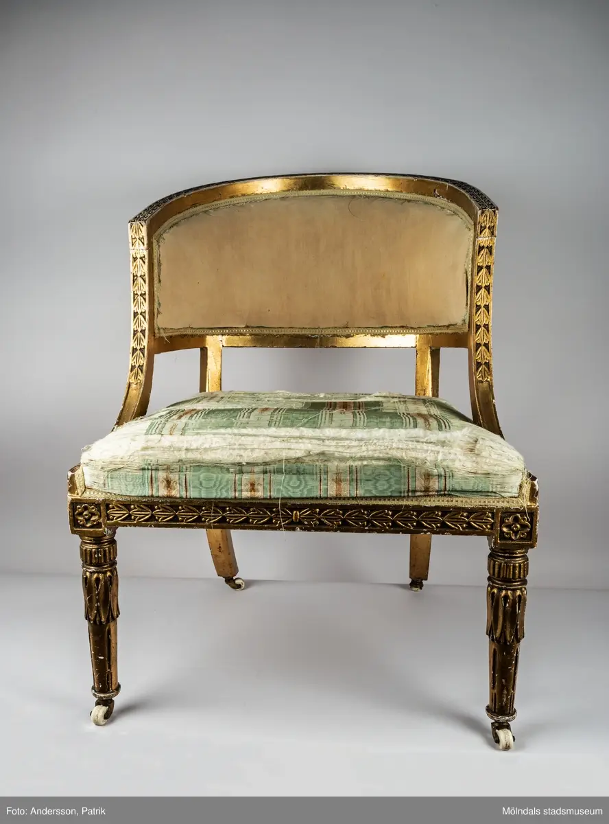 Fåtölj, s.k. "baljfåtölj", sengustaviansk, tillverkad omkring år 1800. 

Kraftigt svängd rygg, raka sarger med skuren dekor, svarvade, bladhölstrade och kannelerade framben, sabelformade bakben. 
Fast stoppad sits och rygg, med sliten, senare klädsel i grönrandigt siden. Bevarad originalstoppning.

Senare bemålning i guld. Äldre rödbrun färg kan anas under förgyllningen.
Benen har kortats av och kompletterats med trissor (hjul) i mässing under 1800-talets andra hälft.

Sitthöjd 440 mm, Bredd 610 mm.
Material: Björk, Tageltyg, Sadelgjord.

Fåtöljen är en av ett par. Båda är signerade med brännstämpel AIS på den bakre sargen. Det finns fyra kända snickare i Lindome som använde signaturen AIS – Anders Johansson i Hassungared (1780-1847), Anders Jonsson i Ingemantorp (1770-1832), Anders Jönsson i Gastorp (1778-1858) och Andreas Johansson i Skår (1793-1871).

Inköpt på Kungsbacka Auktionskammare.
Samhör med MM05442