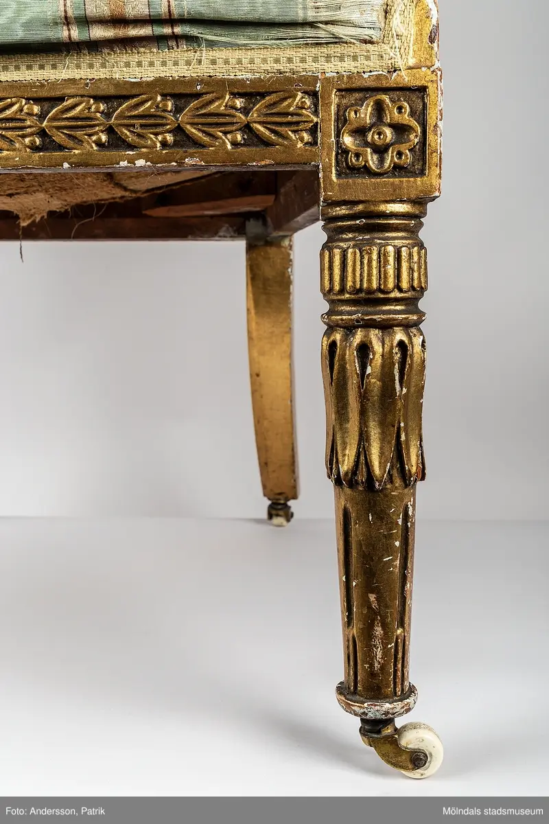 Fåtölj, s.k. "baljfåtölj", sengustaviansk, tillverkad omkring år 1800. 

Kraftigt svängd rygg, raka sarger med skuren dekor, svarvade, bladhölstrade och kannelerade framben, sabelformade bakben. 
Fast stoppad sits och rygg, med sliten, senare klädsel i grönrandigt siden. Bevarad originalstoppning.

Senare bemålning i guld. Äldre rödbrun färg kan anas under förgyllningen.
Benen har kortats av och kompletterats med trissor (hjul) i mässing under 1800-talets andra hälft.

Sitthöjd 440 mm, Bredd 610 mm.
Material: Björk, Tageltyg, Sadelgjord.

Fåtöljen är en av ett par. Båda är signerade med brännstämpel AIS på den bakre sargen. Det finns fyra kända snickare i Lindome som använde signaturen AIS – Anders Johansson i Hassungared (1780-1847), Anders Jonsson i Ingemantorp (1770-1832), Anders Jönsson i Gastorp (1778-1858) och Andreas Johansson i Skår (1793-1871).

Inköpt på Kungsbacka Auktionskammare.
Samhör med MM05442