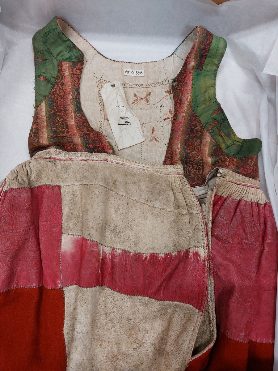 Drakt bestående av liv og stakk som henger sammen. Deler av stakken er av lær/skinn. Den øverste delen av stakken er rødfarget skinn som er stripet hele veien med unntak av et stykke midt foran. Deler av drakta er fôret med ullstoff. Noe av fôret er også av grovt lerrevtsvevd stoff, trolig lin. Den har hekte av metall i sida og på midten i livet. Inni er det en lapp som det står N 69 på. Stakken har ei lomme sydd inn i skinndelen på hyre side, fòret med lin. Hele stakken er fòret. Livet er sydd med faldesting med prikk og har broderier på ryggen. Livet går til rett under bysten.

Fra revisjon: Skinnet er saueskinnspels med pelsen på innsiden. (A.S)