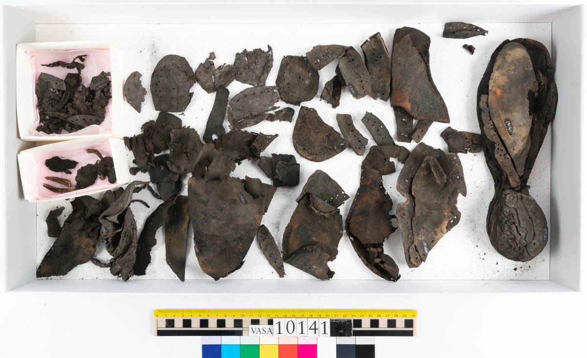 Ett 70-tal delar och fragment av sko: del av skobotten, inlägg, sulor, ovanläder, sidostycken m.m. samt tre fragment textilband samt ett fragment av filt eller tuskaftsväv.

Skobotten, troligtvis från en höger sko. Klacken är uppbyggd av fem klacklappar samt inlägg. Klacken är ritsad på undersidan. Skobottnen har kvarsittande skopligg.

På delarna och fragmenten finns hål efter skopligg och tråd. Lädret är mycket sprött samt har mineral- och rostliknande utfällningar.