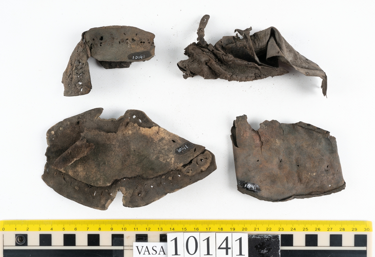 Ett 70-tal delar och fragment av sko: del av skobotten, inlägg, sulor, ovanläder, sidostycken m.m. samt tre fragment textilband samt ett fragment av filt eller tuskaftsväv.

Skobotten, troligtvis från en höger sko. Klacken är uppbyggd av fem klacklappar samt inlägg. Klacken är ritsad på undersidan. Skobottnen har kvarsittande skopligg.

På delarna och fragmenten finns hål efter skopligg och tråd. Lädret är mycket sprött samt har mineral- och rostliknande utfällningar.