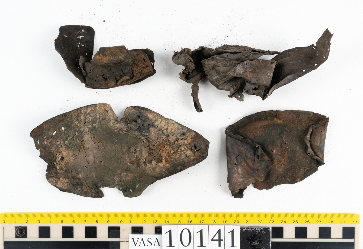 Ett 70-tal delar och fragment av sko: del av skobotten, inlägg, sulor, ovanläder, sidostycken m.m. samt tre fragment textilband samt ett fragment av filt eller tuskaftsväv.

Skobotten, troligtvis från en höger sko. Klacken är uppbyggd av fem klacklappar samt inlägg. Klacken är ritsad på undersidan. Skobottnen har kvarsittande skopligg.

På delarna och fragmenten finns hål efter skopligg och tråd. Lädret är mycket sprött samt har mineral- och rostliknande utfällningar.