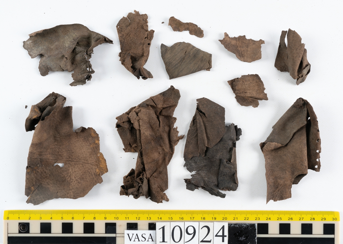 Delar och fragment av stövel.
Ett fragmentariskt stövelskaft. Skaftet är monterat på svart kartong av konserveringsenheten. På skaftets nedre del löper stygnshål. Lädret är mycket fragmentariskt och sprött.
Ett 100-tal större och mindre fragment. På några av fragmenten syns hål efter träpligg och stygn.
Lädret är sprött, tunt och fragmentariskt samt har rostliknande utfällningar.