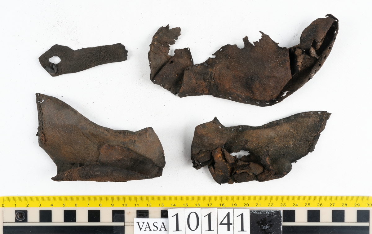 Ett 70-tal delar och fragment av sko: del av skobotten, inlägg, sulor, ovanläder, sidostycken m.m. samt tre fragment textilband samt ett fragment av filt eller tuskaftsväv.

Skobotten, troligtvis från en höger sko. Klacken är uppbyggd av fem klacklappar samt inlägg. Klacken är ritsad på undersidan. Skobottnen har kvarsittande skopligg.

På delarna och fragmenten finns hål efter skopligg och tråd. Lädret är mycket sprött samt har mineral- och rostliknande utfällningar.