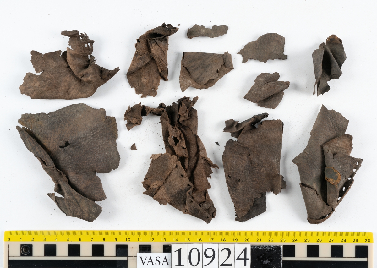 Delar och fragment av stövel.
Ett fragmentariskt stövelskaft. Skaftet är monterat på svart kartong av konserveringsenheten. På skaftets nedre del löper stygnshål. Lädret är mycket fragmentariskt och sprött.
Ett 100-tal större och mindre fragment. På några av fragmenten syns hål efter träpligg och stygn.
Lädret är sprött, tunt och fragmentariskt samt har rostliknande utfällningar.