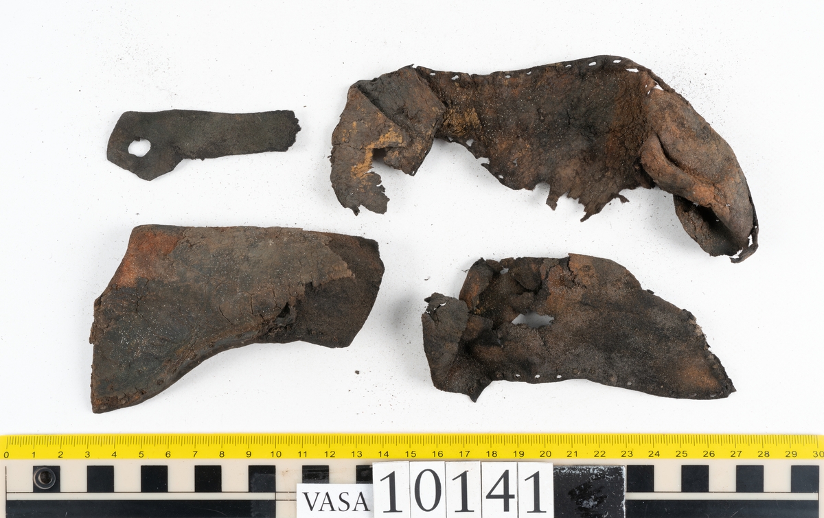 Ett 70-tal delar och fragment av sko: del av skobotten, inlägg, sulor, ovanläder, sidostycken m.m. samt tre fragment textilband samt ett fragment av filt eller tuskaftsväv.

Skobotten, troligtvis från en höger sko. Klacken är uppbyggd av fem klacklappar samt inlägg. Klacken är ritsad på undersidan. Skobottnen har kvarsittande skopligg.

På delarna och fragmenten finns hål efter skopligg och tråd. Lädret är mycket sprött samt har mineral- och rostliknande utfällningar.