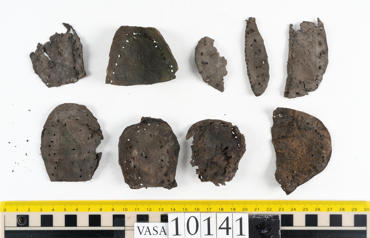 Ett 70-tal delar och fragment av sko: del av skobotten, inlägg, sulor, ovanläder, sidostycken m.m. samt tre fragment textilband samt ett fragment av filt eller tuskaftsväv.

Skobotten, troligtvis från en höger sko. Klacken är uppbyggd av fem klacklappar samt inlägg. Klacken är ritsad på undersidan. Skobottnen har kvarsittande skopligg.

På delarna och fragmenten finns hål efter skopligg och tråd. Lädret är mycket sprött samt har mineral- och rostliknande utfällningar.