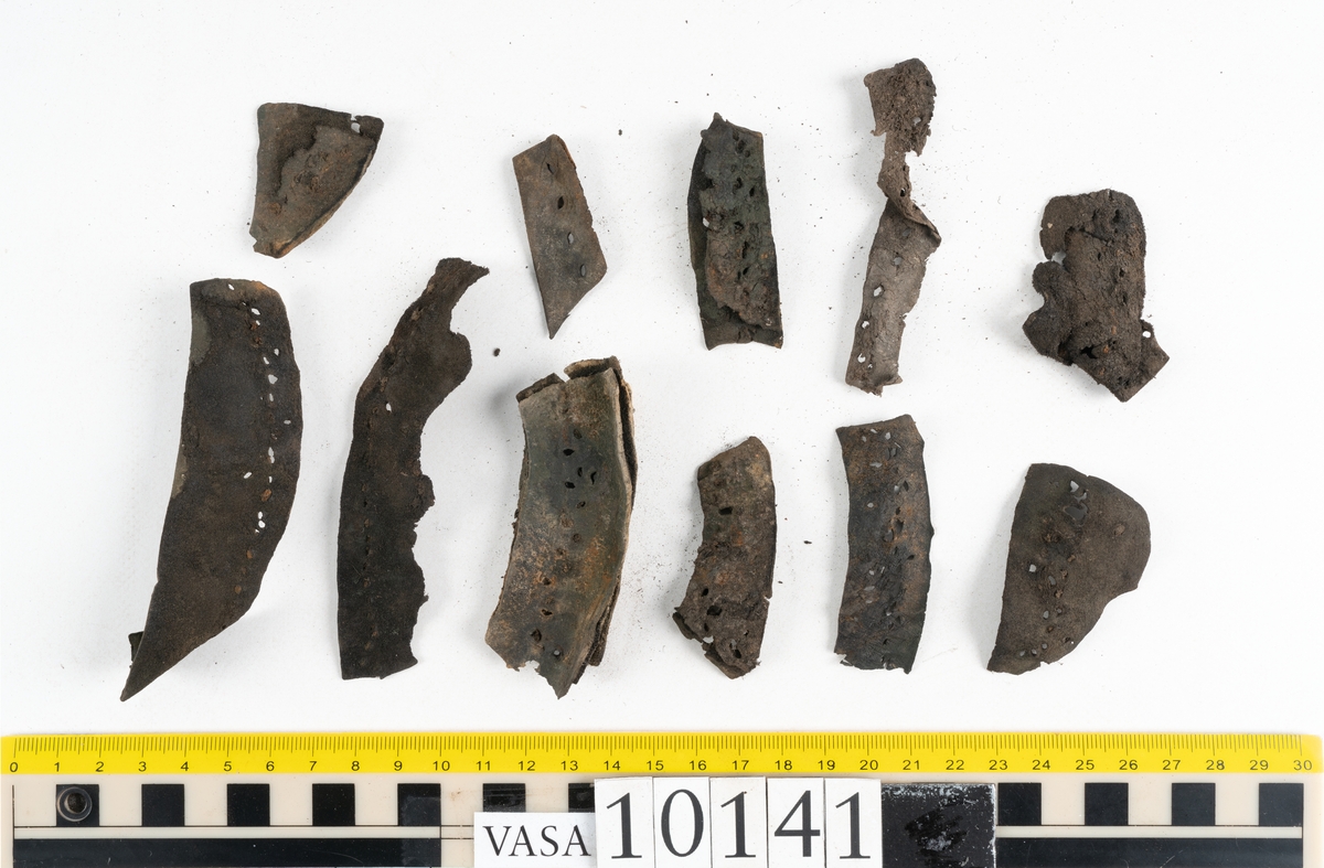 Ett 70-tal delar och fragment av sko: del av skobotten, inlägg, sulor, ovanläder, sidostycken m.m. samt tre fragment textilband samt ett fragment av filt eller tuskaftsväv.

Skobotten, troligtvis från en höger sko. Klacken är uppbyggd av fem klacklappar samt inlägg. Klacken är ritsad på undersidan. Skobottnen har kvarsittande skopligg.

På delarna och fragmenten finns hål efter skopligg och tråd. Lädret är mycket sprött samt har mineral- och rostliknande utfällningar.