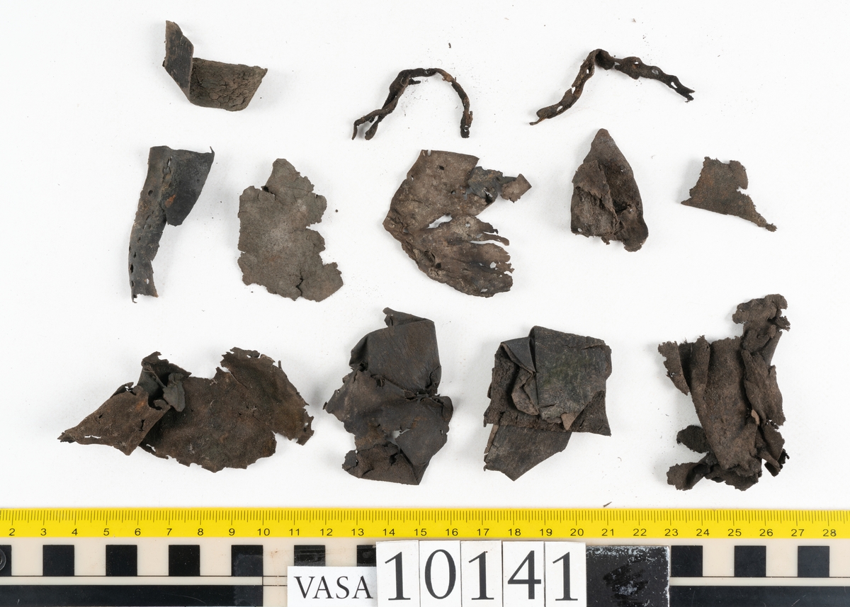 Ett 70-tal delar och fragment av sko: del av skobotten, inlägg, sulor, ovanläder, sidostycken m.m. samt tre fragment textilband samt ett fragment av filt eller tuskaftsväv.

Skobotten, troligtvis från en höger sko. Klacken är uppbyggd av fem klacklappar samt inlägg. Klacken är ritsad på undersidan. Skobottnen har kvarsittande skopligg.

På delarna och fragmenten finns hål efter skopligg och tråd. Lädret är mycket sprött samt har mineral- och rostliknande utfällningar.