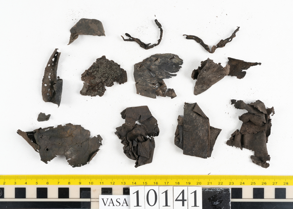 Ett 70-tal delar och fragment av sko: del av skobotten, inlägg, sulor, ovanläder, sidostycken m.m. samt tre fragment textilband samt ett fragment av filt eller tuskaftsväv.

Skobotten, troligtvis från en höger sko. Klacken är uppbyggd av fem klacklappar samt inlägg. Klacken är ritsad på undersidan. Skobottnen har kvarsittande skopligg.

På delarna och fragmenten finns hål efter skopligg och tråd. Lädret är mycket sprött samt har mineral- och rostliknande utfällningar.