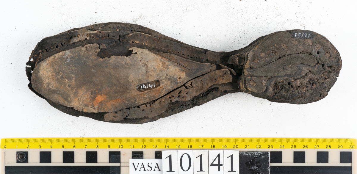 Ett 70-tal delar och fragment av sko: del av skobotten, inlägg, sulor, ovanläder, sidostycken m.m. samt tre fragment textilband samt ett fragment av filt eller tuskaftsväv.

Skobotten, troligtvis från en höger sko. Klacken är uppbyggd av fem klacklappar samt inlägg. Klacken är ritsad på undersidan. Skobottnen har kvarsittande skopligg.

På delarna och fragmenten finns hål efter skopligg och tråd. Lädret är mycket sprött samt har mineral- och rostliknande utfällningar.