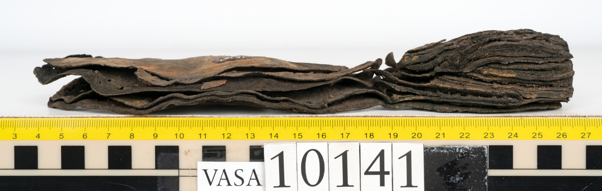 Ett 70-tal delar och fragment av sko: del av skobotten, inlägg, sulor, ovanläder, sidostycken m.m. samt tre fragment textilband samt ett fragment av filt eller tuskaftsväv.

Skobotten, troligtvis från en höger sko. Klacken är uppbyggd av fem klacklappar samt inlägg. Klacken är ritsad på undersidan. Skobottnen har kvarsittande skopligg.

På delarna och fragmenten finns hål efter skopligg och tråd. Lädret är mycket sprött samt har mineral- och rostliknande utfällningar.