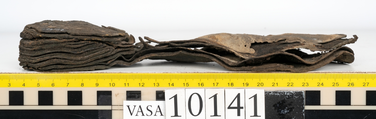Ett 70-tal delar och fragment av sko: del av skobotten, inlägg, sulor, ovanläder, sidostycken m.m. samt tre fragment textilband samt ett fragment av filt eller tuskaftsväv.

Skobotten, troligtvis från en höger sko. Klacken är uppbyggd av fem klacklappar samt inlägg. Klacken är ritsad på undersidan. Skobottnen har kvarsittande skopligg.

På delarna och fragmenten finns hål efter skopligg och tråd. Lädret är mycket sprött samt har mineral- och rostliknande utfällningar.