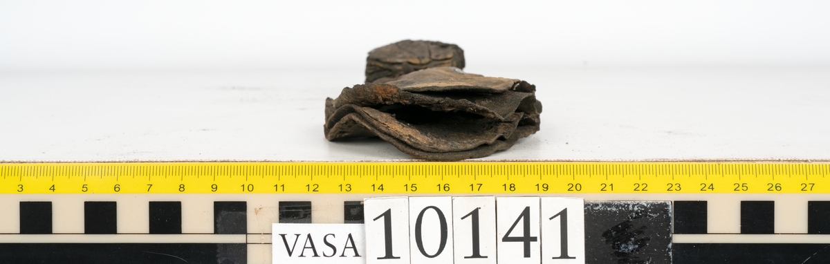 Ett 70-tal delar och fragment av sko: del av skobotten, inlägg, sulor, ovanläder, sidostycken m.m. samt tre fragment textilband samt ett fragment av filt eller tuskaftsväv.

Skobotten, troligtvis från en höger sko. Klacken är uppbyggd av fem klacklappar samt inlägg. Klacken är ritsad på undersidan. Skobottnen har kvarsittande skopligg.

På delarna och fragmenten finns hål efter skopligg och tråd. Lädret är mycket sprött samt har mineral- och rostliknande utfällningar.