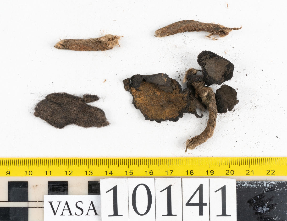 Ett 70-tal delar och fragment av sko: del av skobotten, inlägg, sulor, ovanläder, sidostycken m.m. samt tre fragment textilband samt ett fragment av filt eller tuskaftsväv.

Skobotten, troligtvis från en höger sko. Klacken är uppbyggd av fem klacklappar samt inlägg. Klacken är ritsad på undersidan. Skobottnen har kvarsittande skopligg.

På delarna och fragmenten finns hål efter skopligg och tråd. Lädret är mycket sprött samt har mineral- och rostliknande utfällningar.