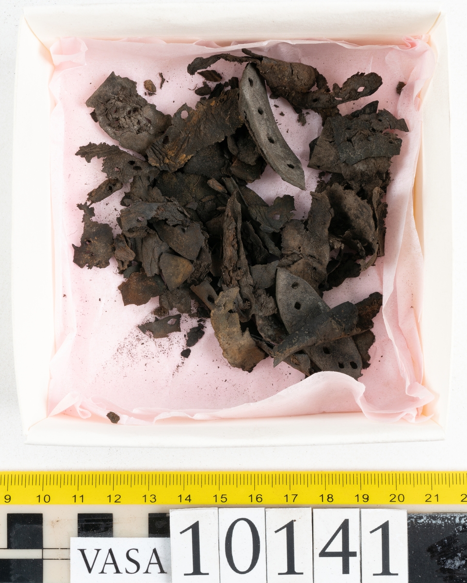 Ett 70-tal delar och fragment av sko: del av skobotten, inlägg, sulor, ovanläder, sidostycken m.m. samt tre fragment textilband samt ett fragment av filt eller tuskaftsväv.

Skobotten, troligtvis från en höger sko. Klacken är uppbyggd av fem klacklappar samt inlägg. Klacken är ritsad på undersidan. Skobottnen har kvarsittande skopligg.

På delarna och fragmenten finns hål efter skopligg och tråd. Lädret är mycket sprött samt har mineral- och rostliknande utfällningar.