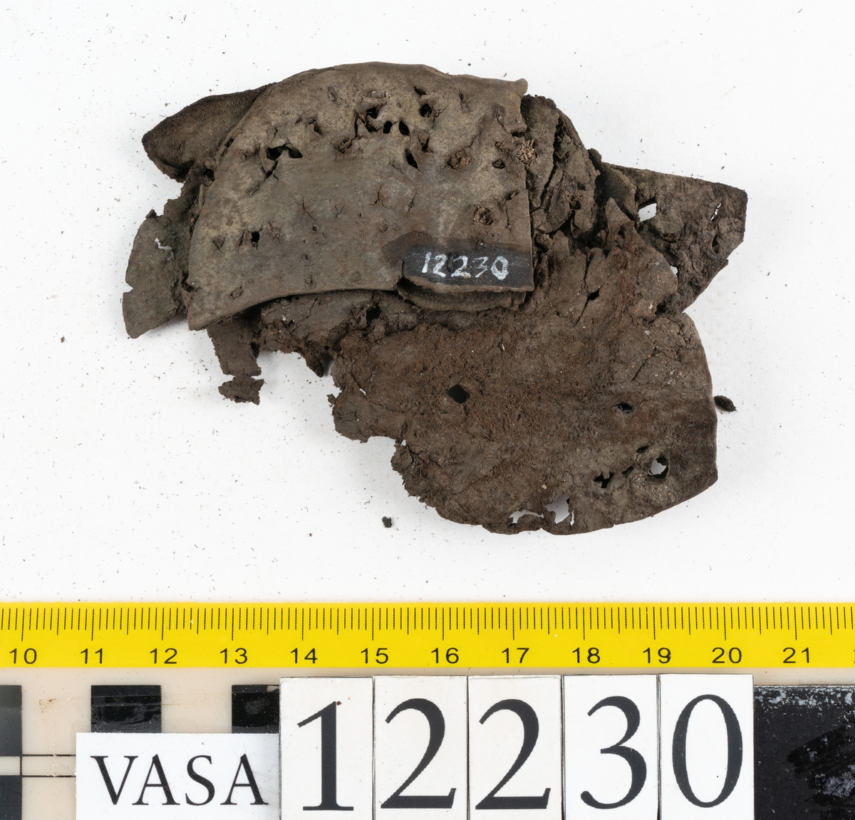 Delar och fragment från sko. Totalt rör det sig om ett 40-tal delar och fragment från ovanläder, sulor, klack och inlägg.
En träklack, fyndnummer 12230. Klacken sidor är skurna snett. På undersidan finns kvarsittande träpligg samt hål efter träpligg. Klacken är något snedsliten. Träet är fuktigt.
Delar av två skobottnar, fyndnummer 12230:1 samt 12230:2. Bottnarna härrör från ett och samma par skor. Skobottnarna har kvarsittande träpligg och tråd. Skobottennarna är delvist fragmentariska och fyndnummer 12230 saknar hälpartiet. Lädret är relativt kraftigt men sprött.